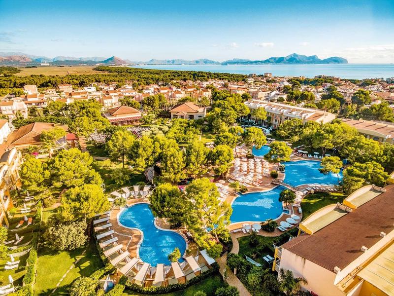 Große Poolanlage mit geschwungenen Formen, umgeben von grünen Bäumen und Sonnenschirmen in einer Ferienanlage.