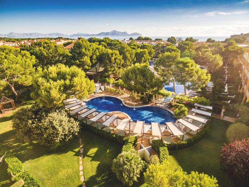 Rundlicher Pool mit Liegen, umgeben von Bäumen und viel grüner Vegetation unter blauem Himmel.