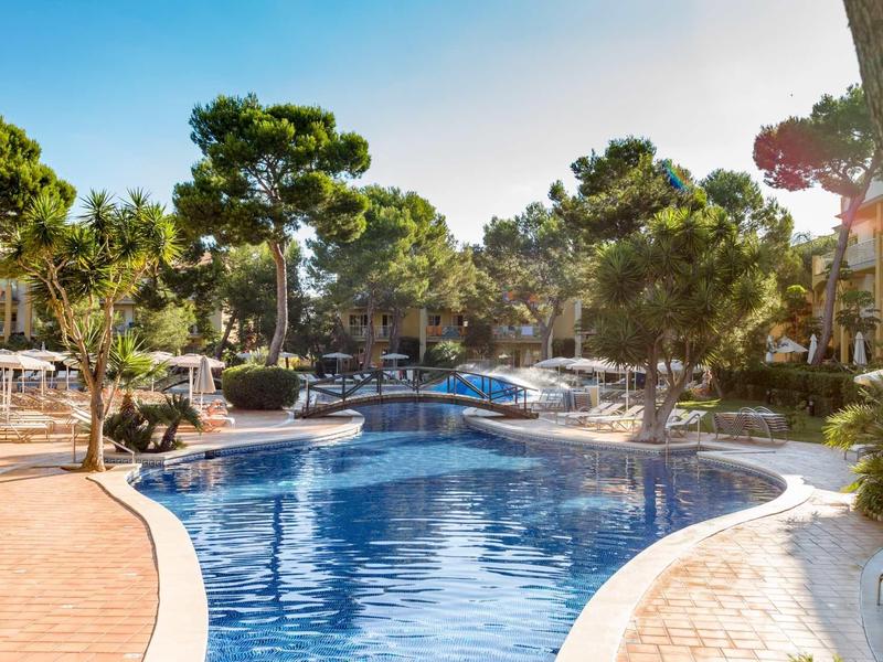 Großer Pool mit blauer Wasseroberfläche, umgeben von Bäumen, Liegestühlen und Sonnenschirmen.