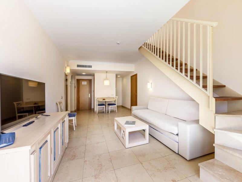 Helles Wohnzimmer mit beigem Sofa, Treppe rechts, Fernseher links und Essbereich im Hintergrund.