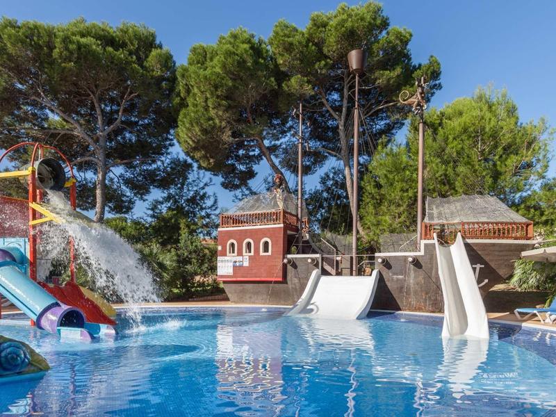 Pool mit großen Wasserrutschen, Wasserspiel und Bäumen im Hintergrund bei klarem Himmel.