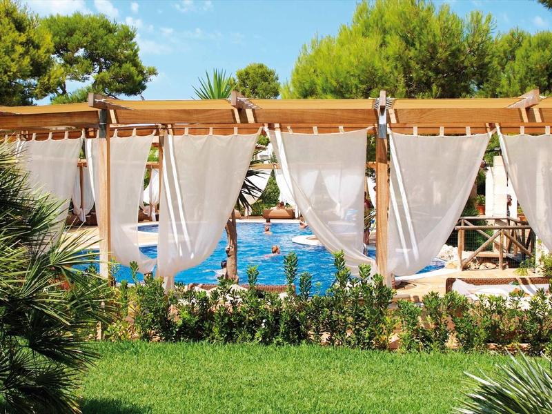 Blaue Poollandschaft mit weißen Vorhängen, Holzpergola und grüner Pflanzenumgebung.