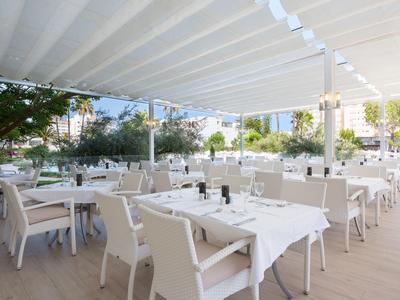 Offenes Restaurant mit weißen Tischen und Stühlen unter einem großen Sonnensegel, viele Bäume im Hintergrund.