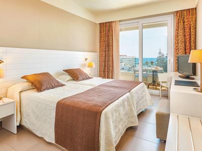 Helles Schlafzimmer mit Doppelbett, zwei Nachttischen, Lampe, Balkon und Vorhängen in Beige- und Brauntönen.