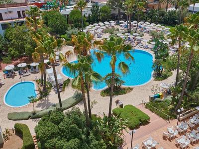 Großer Hotelpool mit Palmen, Liegestühlen und Sonnenschirmen in einer grünen Gartenanlage.