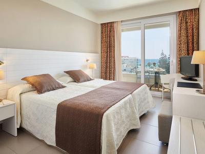 Helles Hotelzimmer mit Doppelbett, Nachttischen, Lampen und Balkon mit Meerblick.