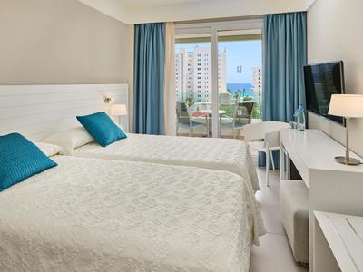 Camera doppia moderna con due letti singoli, cuscini blu, scrivania e vista sulla città e sul mare.