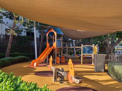 Parco giochi per bambini con scivolo e struttura da arrampicata sotto ombrelloni nel giardino di un hotel.