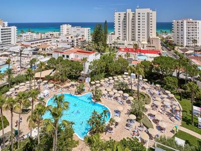 Vista di un hotel con una grande piscina, lettini e il mare blu sullo sfondo.