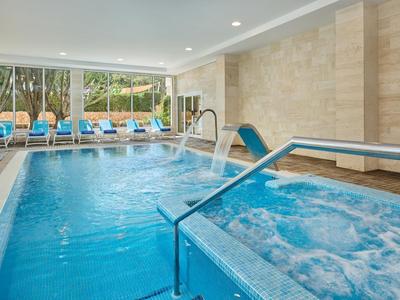 Area piscina interna moderna con vasca idromassaggio, getti d'acqua e sedie a sdraio blu sullo sfondo.
