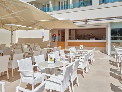 Terrazza moderna con sedie e tavoli bianchi davanti a un hotel, con ombrelloni e bar.
