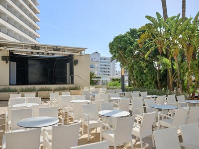 Area esterna di un hotel con tavoli e sedie bianchi e alberi alti sotto un cielo azzurro.