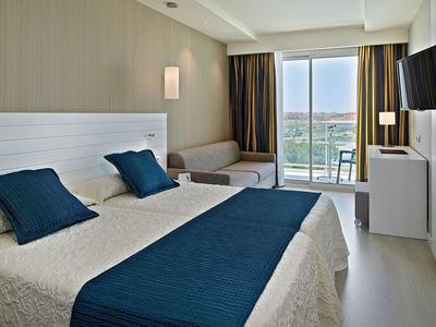 Modernes Hotelzimmer mit Doppelbett, blauen Akzenten, Balkon und Aussicht auf Bäume und Gebäude.