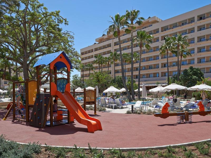 Spielplatz mit roter Rutsche neben Hotelgebäude und Palmen unter blauem Himmel.