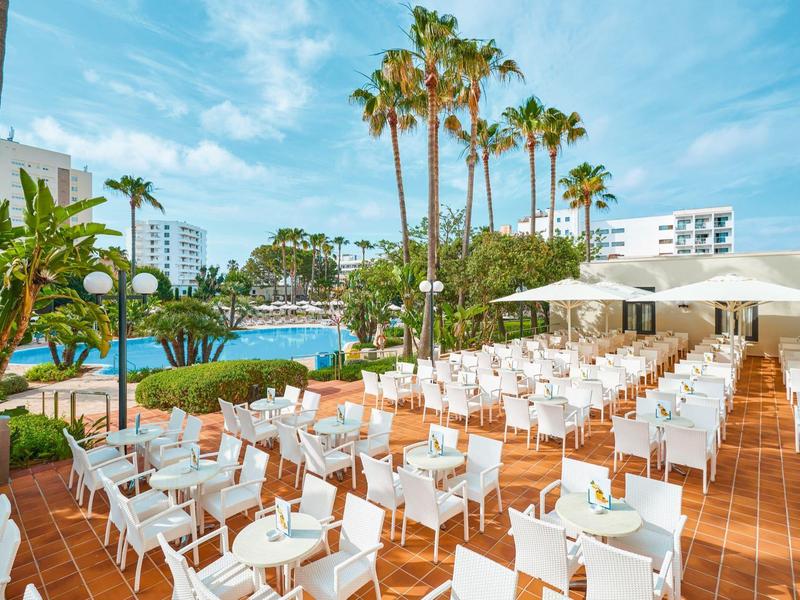 Große Terrasse mit weißen Stühlen und Tischen, Palmen und Pool vor blauem Himmel.