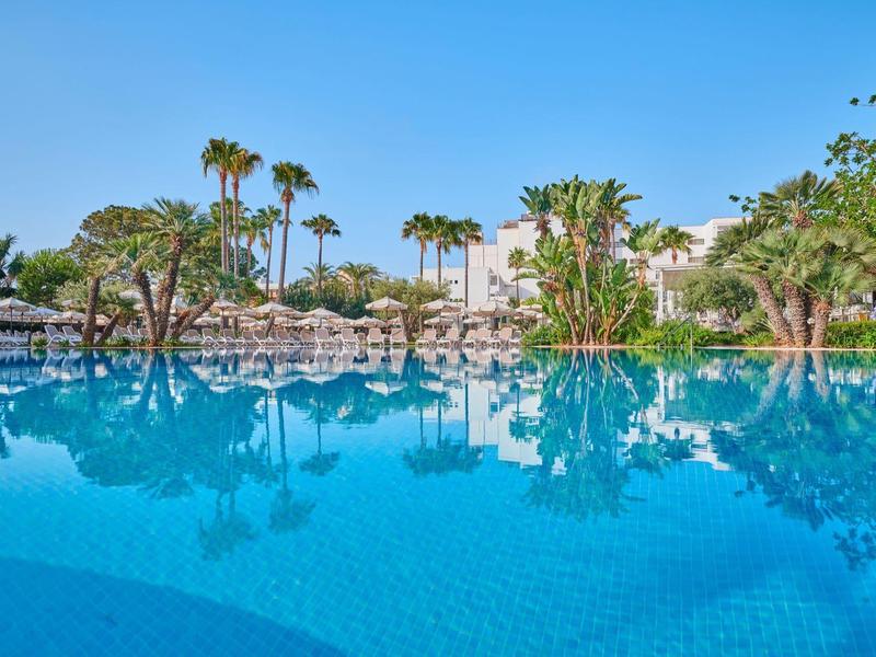 Großer Pool mit Palmen und Sonnenliegen in einem Hotelresort bei klarem Himmel.