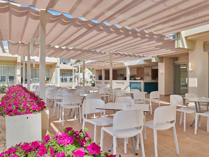 Leerer Außenbereich eines Cafés mit weißen Stühlen und rosa Blumen unter einem Sonnendach.