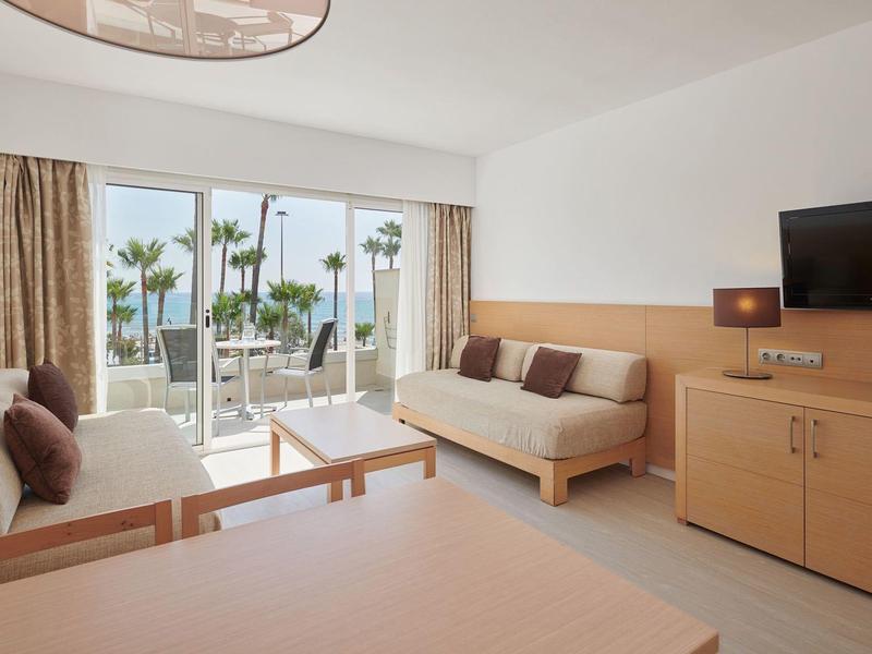 Gemütliches Hotelzimmer mit Meerblick, Sofa, TV und Balkon mit Palmen.