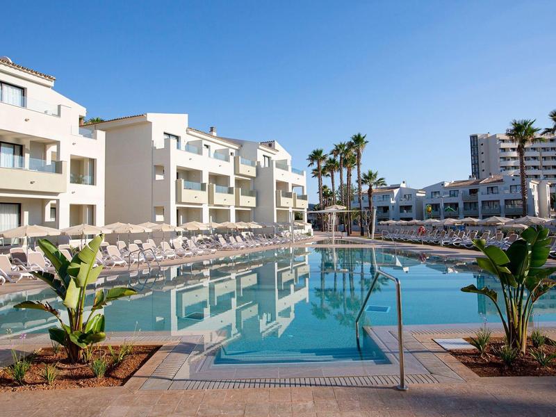 Moderner Hotelpool mit Liegestühlen und tropischen Pflanzen unter klarem Himmel.