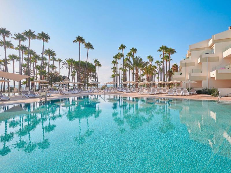 Großer Hotelpool mit zahlreichen Liegestühlen und Palmen bei sonnigem Wetter.