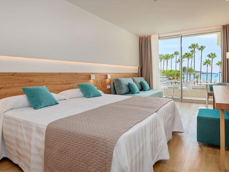 Helles Hotelzimmer mit zwei Betten, türkisfarbenen Kissen und Blick auf Palmen und Meer.