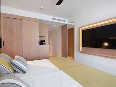 Nowoczesny pokój hotelowy z łóżkiem podwójnym, telewizorem na ścianie i jasną drewnianą szafą.