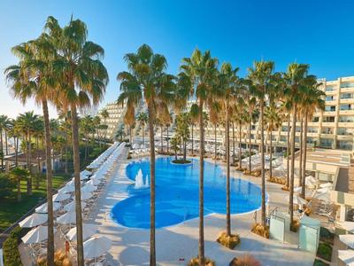 Großer Pool mit blauer Wasserfläche, umgeben von Palmen und weißen Liegestühlen neben mehrstöckigem Hotel.