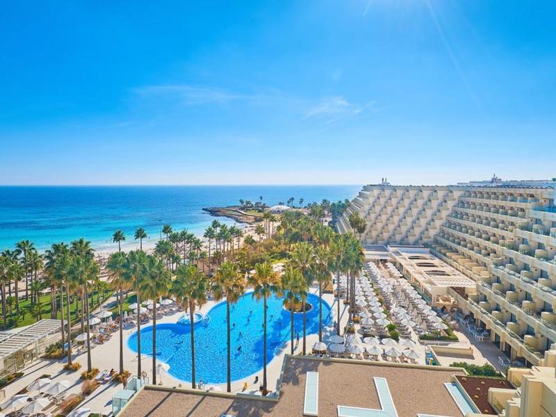 Widok na kompleks hotelowy z basenem, palmami i dostępem do plaży pod błękitnym niebem.