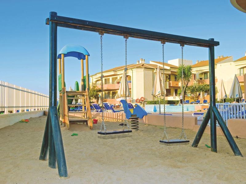 Swing set with multiple seats on sandy ground in front of hotel buildings under clear sky.