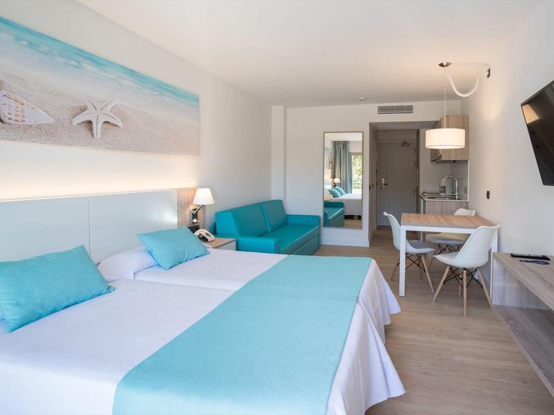 Bright hotel room with double bed, turquoise pillows, sofa, dining table, and wall-mounted TV.