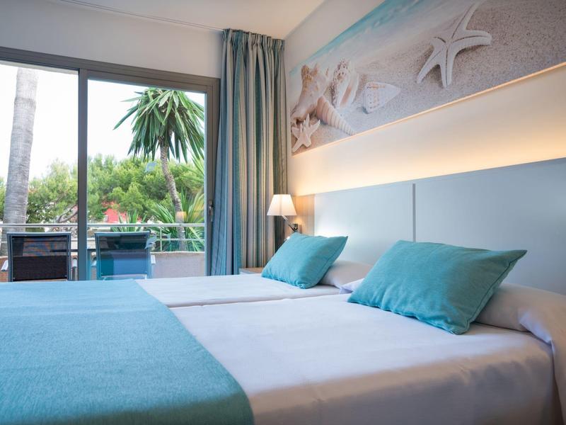 Bright hotel room with two beds, blue pillows, view of terrace and green plants.