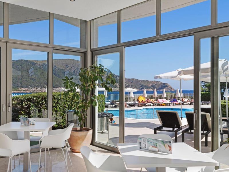 Modern indoor area with white chairs and tables overlooking pool and sea with mountains in the background.