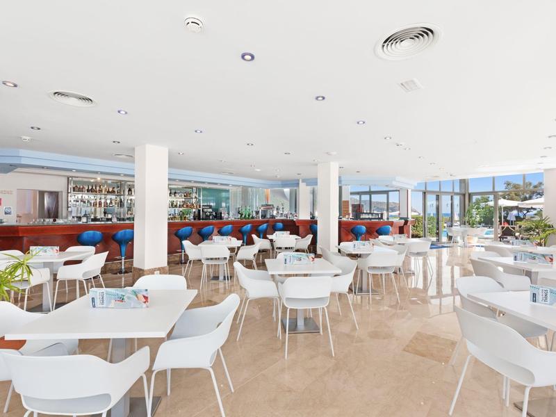 Bright dining area with white tables and chairs and a long bar in the background.