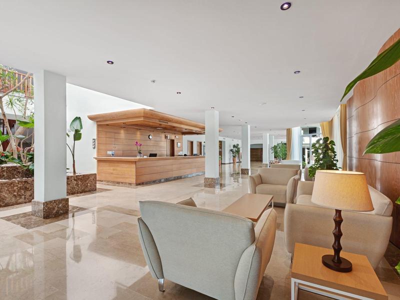 Bright hotel lobby with seating area, wooden reception desk, and large windows showing plants.