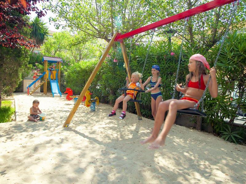 Bambini che giocano sulle altalene in un giardino verde e soleggiato con terreno sabbioso.