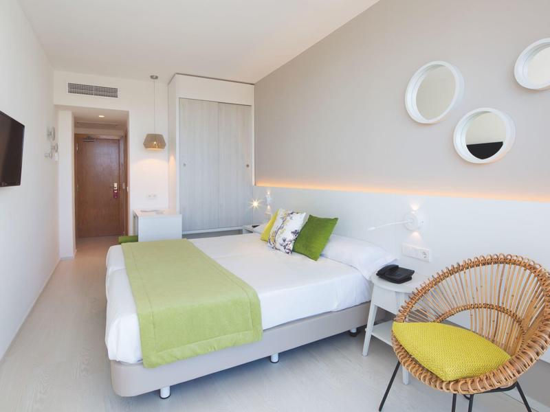 Camera d'hotel moderna con letto matrimoniale, biancheria verde e bianca e sedia in rattan con cuscino giallo.