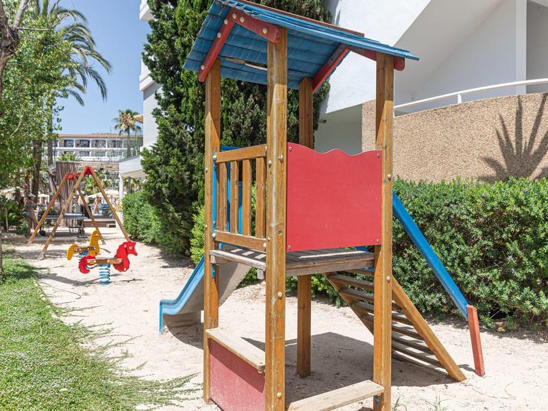 Area giochi per bambini con struttura per arrampicarsi, scivolo e zona sabbiosa accanto a un edificio residenziale.