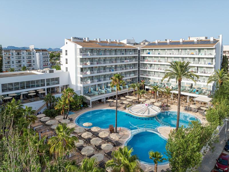 Grande hotel su più piani con piscina e palme nel cortile sotto un cielo azzurro.