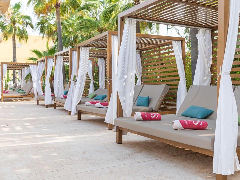 Letti da spiaggia in legno moderni con tende bianche e cuscini colorati sotto le palme.