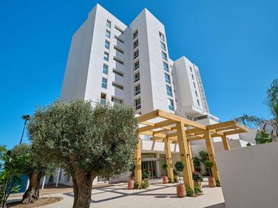 Modern wit hotelgebouw met pergola en olijfbomen onder een heldere blauwe lucht.