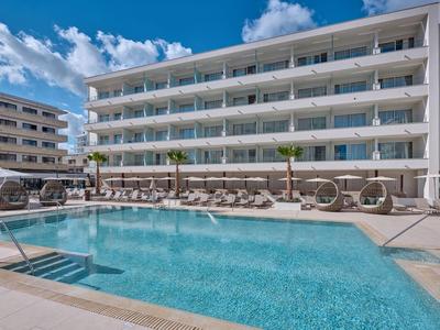 Modern hotel complex with large swimming pool, sun loungers, and clear sky in the background.