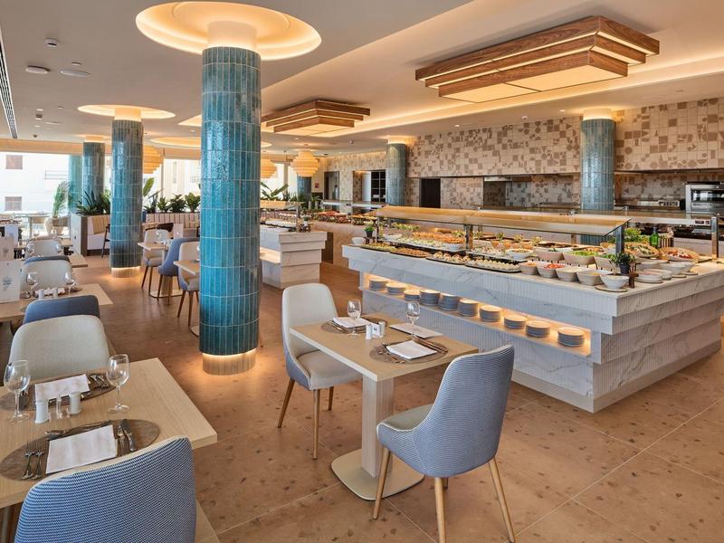 Licht modern hotelrestaurant met blauwe zuilen, grijze stoelen en buffetgebied met stenen muur.