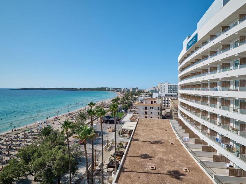 Hotel met balkonkamers naast strand en zee onder heldere blauwe lucht.