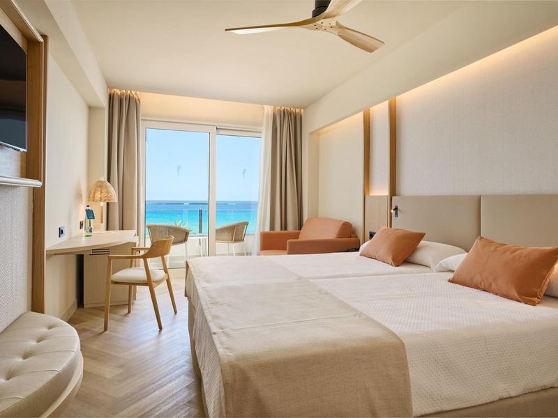 Lichte hotelkamer met twee bedden, zithoek en zeezicht door grote ramen.