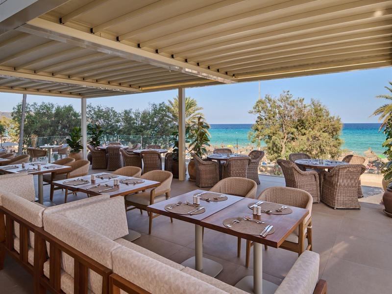 Overdekt terras met tafels, stoelen en uitzicht op zee en bomen.
