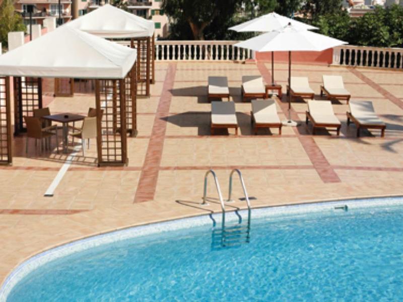 Zona de piscina vacía con tumbonas y sombrillas sobre terraza pavimentada a plena luz del día.