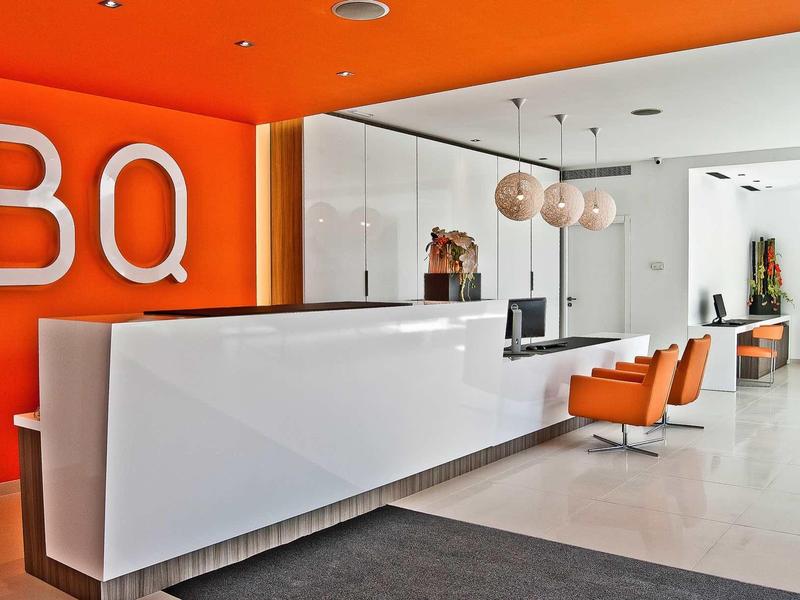 Recepción moderna de hotel con mostrador blanco y sillas naranjas frente a pared naranja con logo BQ.
