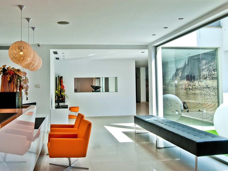 Área de salón de hotel moderno y luminoso con sillones naranjas, bancos largos y grandes ventanales.