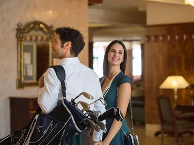 Couple avec sacs de golf dans un hall d'hôtel élégant, tous deux bien habillés, femme souriante.