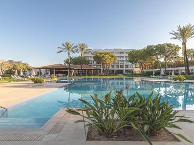 Hotel met een groot zwembad omgeven door parasols en palmbomen onder een heldere lucht.